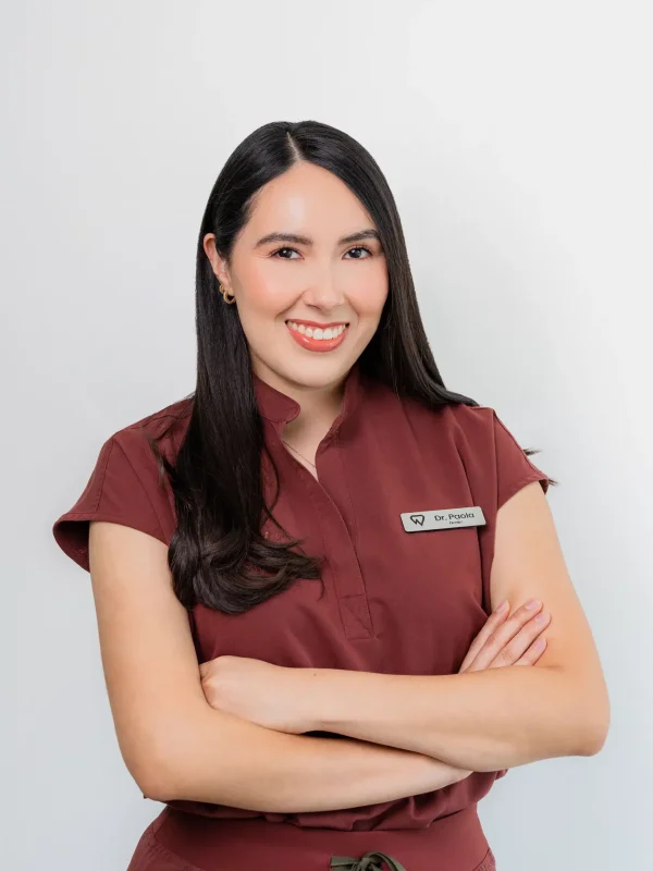 Woman with long dark hair wearing a maroon uniform and name tag stands against a plain background with arms crossed, smiling at the camera.