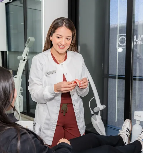 A dentist in a white coat shows a dental model to a patient seated in a dental chair, explaining the root canal procedure in Tijuana inside a modern clinic with large windows.